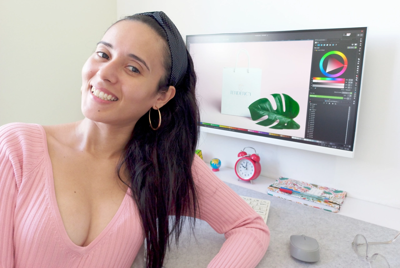 Foto de pefil da Ana Quasar sorrindo em frente ao monitor do seu computador com seus trabalhos como designer abertos na tela